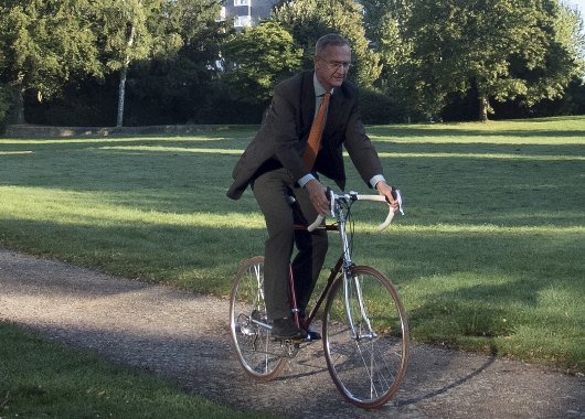 Mann im Anzug fährt Rad über Kiesweg durch einen Park.