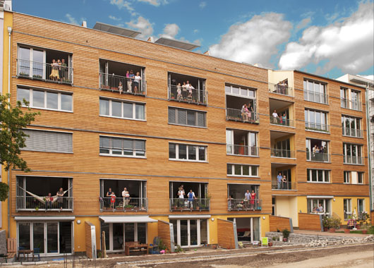 Holzwohnhaus mit Bewohner*innen auf Balkonen.