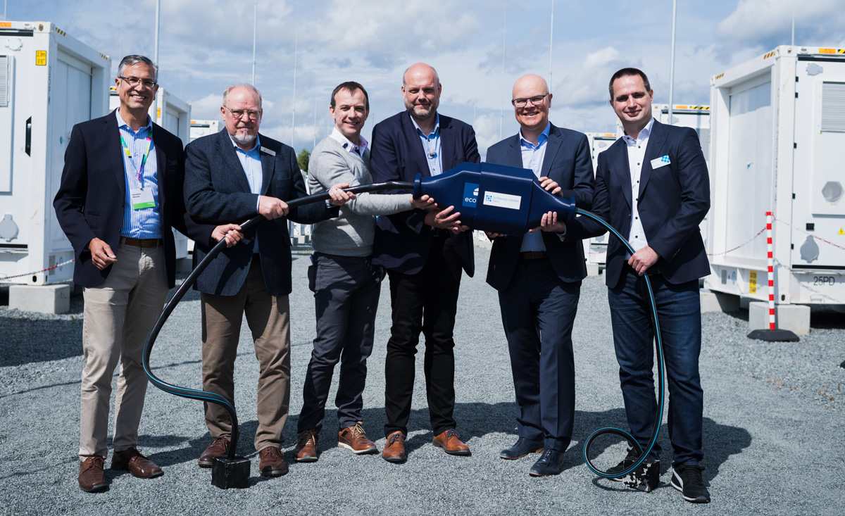 Einweihung des Batteriespeichers von Eco Stor in Bollingstedt, finanziert von der GLS Bank 6 maennlich gelesene Personen halten ein Kabel, im Hintergrund Batteriespeicher