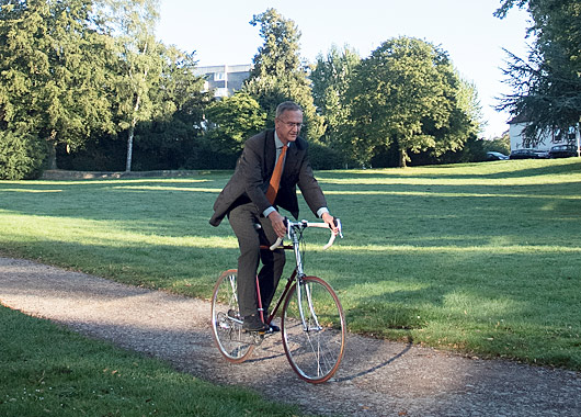 Man im Anzug fährt Rennrad über einen Schotterweg in einem Park.