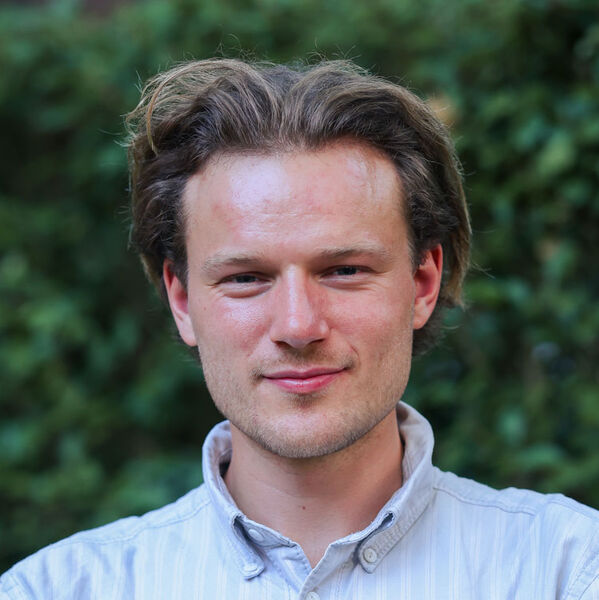 Männlich gelesene Person mit hellbraunem Haar lächelt im Freien vor grünem Laub.