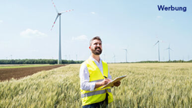 Männlich gelesene Person mit Schreibblock steht in einem Getreidefeld in einem Windpark