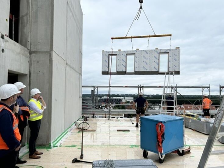 Auf einer Baustelle wird ein Fassadenelement am Kran hochgezogen und schwebt in der Luft. Menschen stehen am Rand und schauen zu.