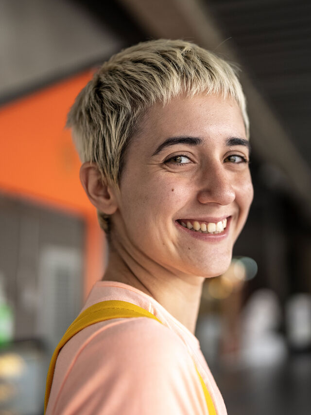 Lächelnde Person mit kurzen blonden Haaren, im Haus, mit rosa Hemd.