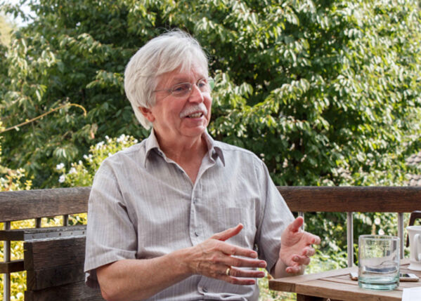 Männlich gelesene ältere Person sitzt am Tisch in grüner Umgebung und erzählt.