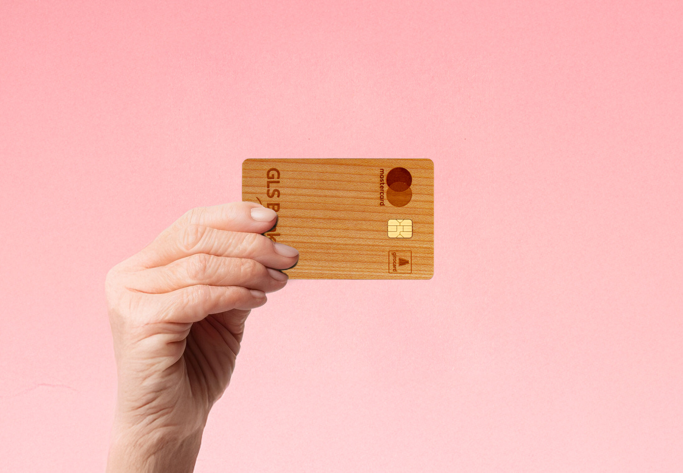 Eine Hand hält eine hölzerne Kreditkarte vor einem Hintergrund mit rosa Farbverlauf.
