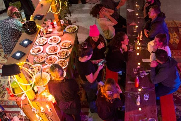 Zwei lange Tafeln mit Essen, weihnachtlicher Beleuchtung und vielen Personen die gemeinsam Essen.