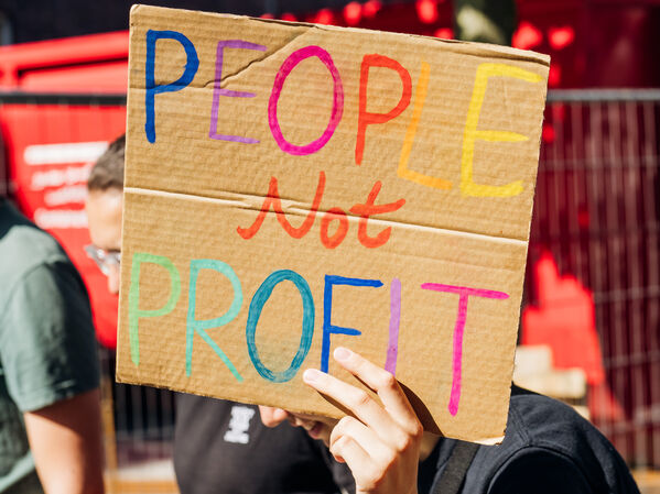 Eine Person hält ein Pappschild, auf dem in bunten Buchstaben "People Not Profit" steht.