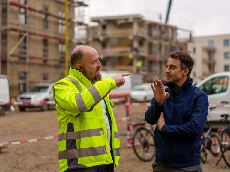 Zwei männlich gelesen Personen unterhalten sich auf einer Baustelle; einer trägt eine gelbe Sicherheitsweste.