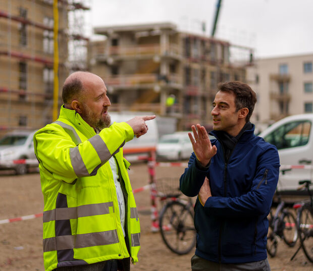 Zwei männlich gelesen Personen unterhalten sich auf einer Baustelle; einer trägt eine gelbe Sicherheitsweste.