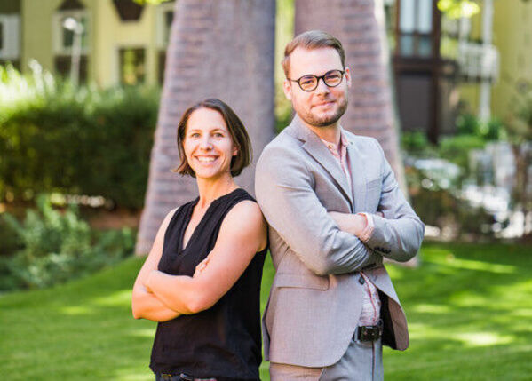 Zwei Personen stehen Rücken an Rücken im Freien, beide lächeln mit verschränkten Armen.