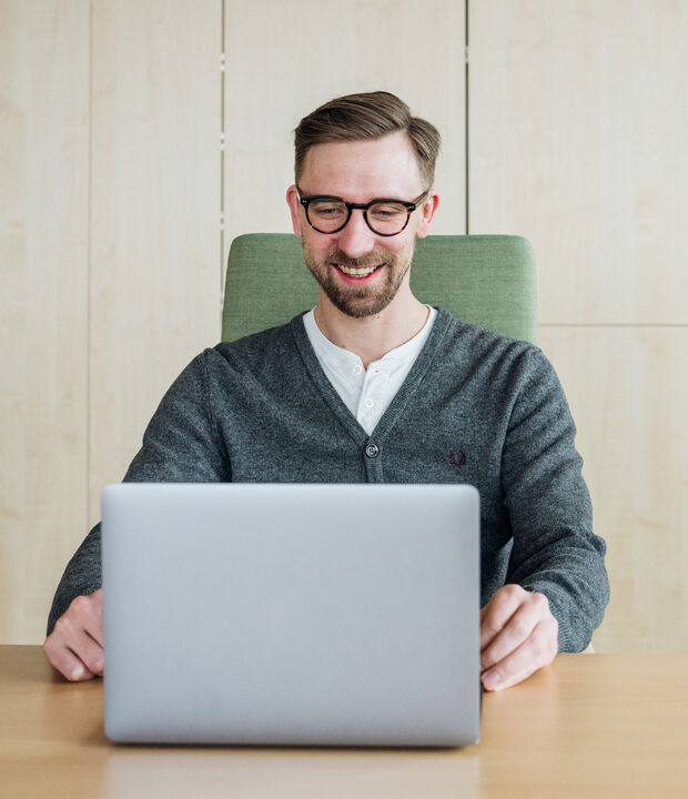 Männlich gelesene Person mit Brille, der einen Laptop an einem Schreibtisch in einem hellen, modernen Büro benutzt.