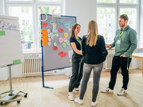 Drei Personen stehen in einem hellen Raum in der Nähe eines Whiteboards und einer Pinnwand und unterhalten sich.