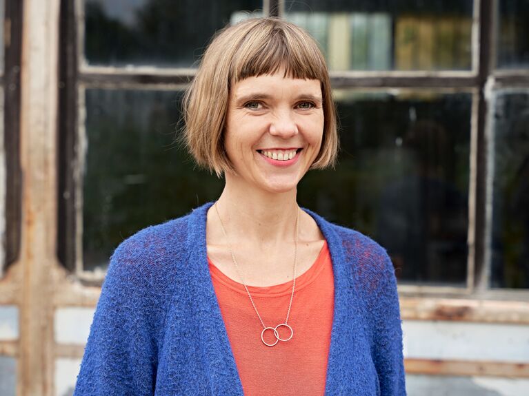 Draußen lächelt eine Frau mit kurzen Haaren, die einen blauen Pullover und ein orangefarbenes Oberteil trägt.