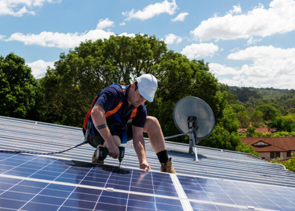 Männlich gelesene Person in Arbeitskleidung installiert eine Solaranlage auf einem Dach.