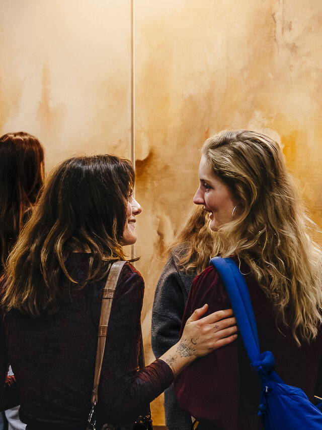 Menschen betrachten abstrakte Kunst in einer Galerie; zwei Frauen unterhalten sich im Vordergrund.