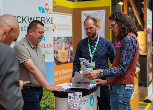 Vier Männer stehen um einen Stand bei einer Geschäfts- oder Informationsveranstaltung in einem Gebäude.