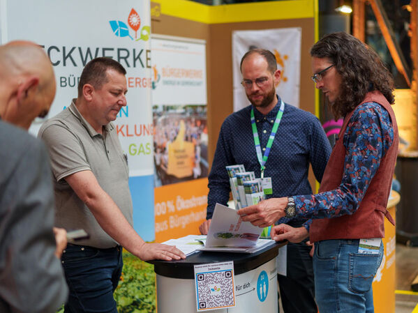 Vier Männer stehen um einen Stand bei einer Geschäfts- oder Informationsveranstaltung in einem Gebäude.