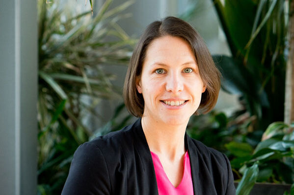 Eine Frau mit kurzen braunen Haaren lächelt, sie trägt eine schwarze Jacke und ein rosa Oberteil.