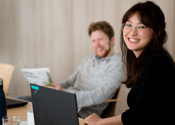 Frau lächelt am Schreibtisch mit Laptop, Mann im Hintergrund hält eine Zeitung und lacht.