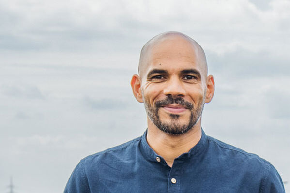 Ein Mann mit rasiertem Kopf und Bart lächelt im Freien in einem blauen Hemd.