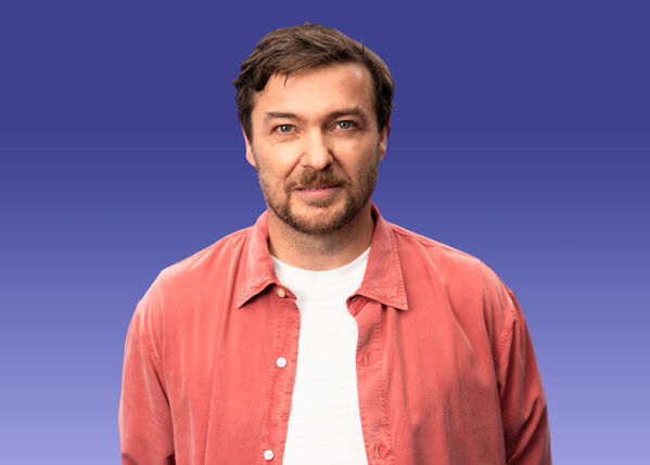 Mann mit braunen Haaren und Bart in einem korallenroten Hemd auf einem blauen Hintergrund mit Farbverlauf.