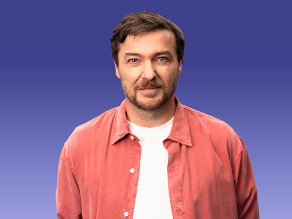 Mann mit braunen Haaren und Bart in einem korallenroten Hemd auf einem blauen Hintergrund mit Farbverlauf.