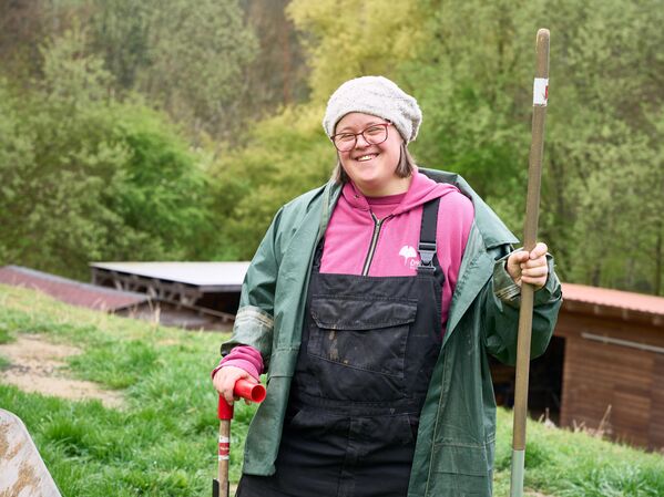 Weiblich gelesene Person mit Mütze und Parka hält einen Stiel steht vor einer Wiese und lächelt