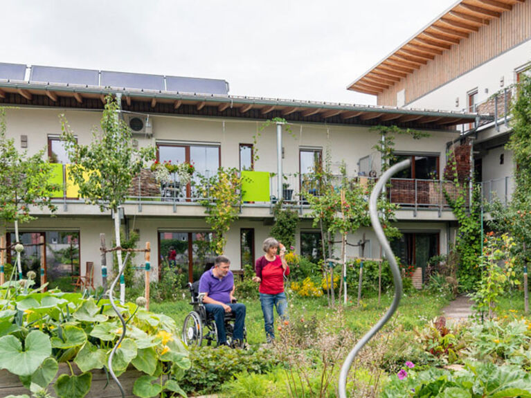 Mensch im Rollstuhl mit Begleitperson in einem Bauerngarten, im Hintergrund ein Wohngebäude