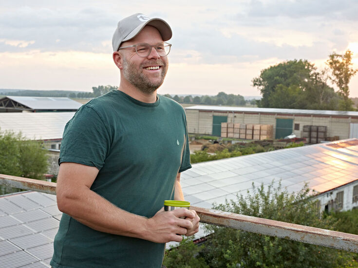 Mann mit Brille und Mütze, lächelnd, mit einer Tasse in der Hand, steht auf einem Dach mit Sonnenkollektoren.