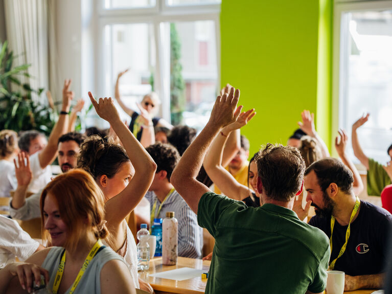 Viele Personen sitzen an zwei langen Tischen und melden sich.