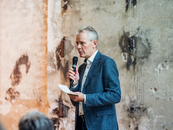 Ein Mann im Anzug spricht in ein Mikrofon und hält Papiere an eine abgenutzte Wand.