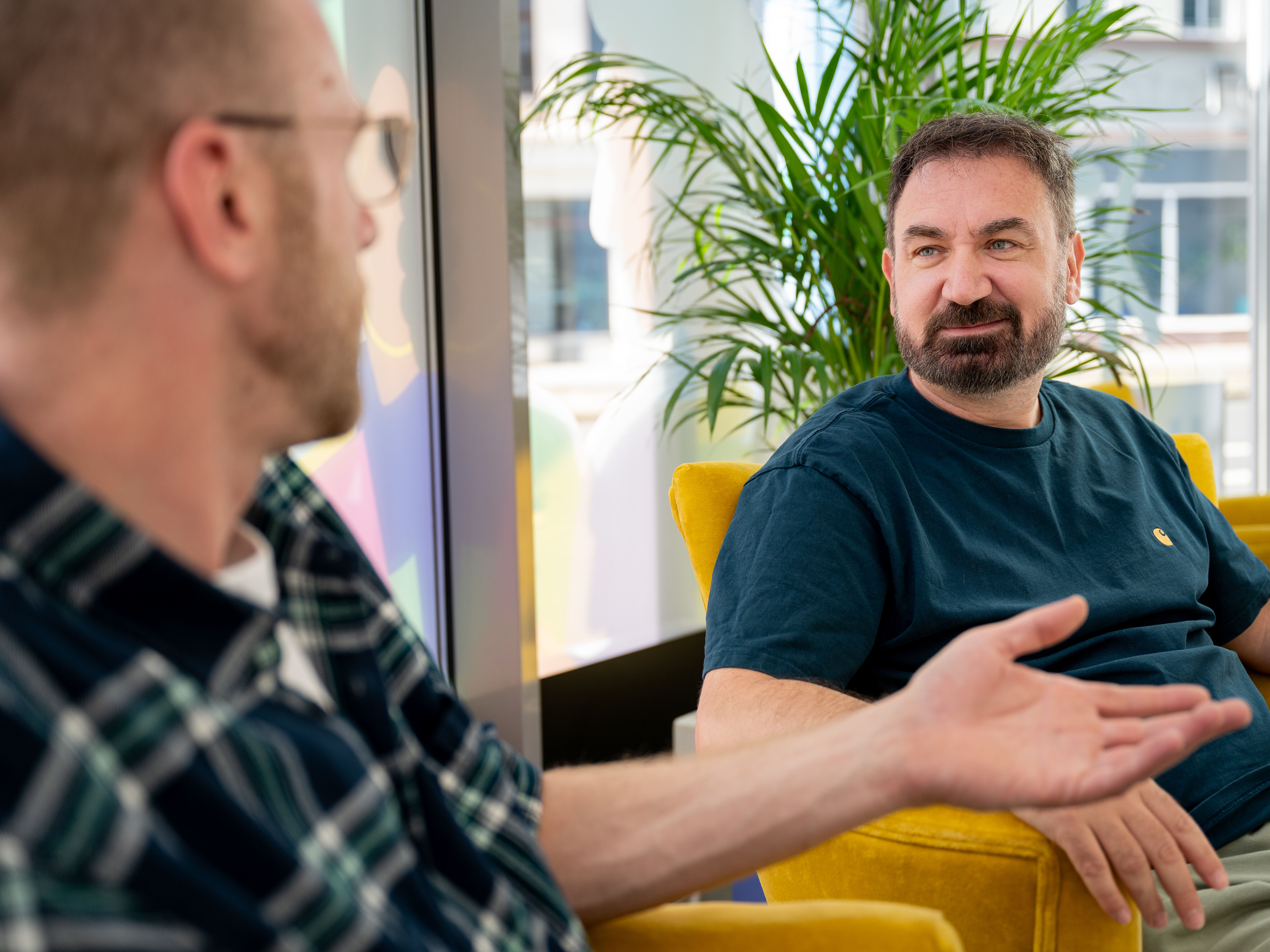 Zwei Männer sitzen auf gelben Stühlen in einem Haus und unterhalten sich in der Nähe eines Fensters.