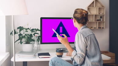 Frau schaut auf einen Computerbildschirm mit einem Warnsymbol, während sie ein Telefon hält.