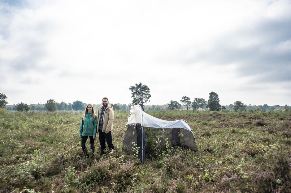 Zwei Personen stehen neben einem Netzzelt auf einem grasbewachsenen, offenen Feld unter einem bewölkten Himmel.