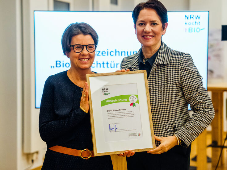 Zwei Frauen stehen in einem Haus, halten eine gerahmte Urkunde in der Hand und lächeln in die Kamera.