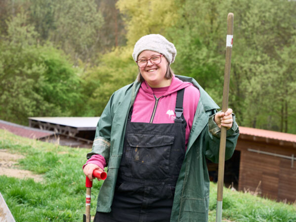 Weiblich gelesene Person steht mit Gartenwerkzeug im Grünen. 