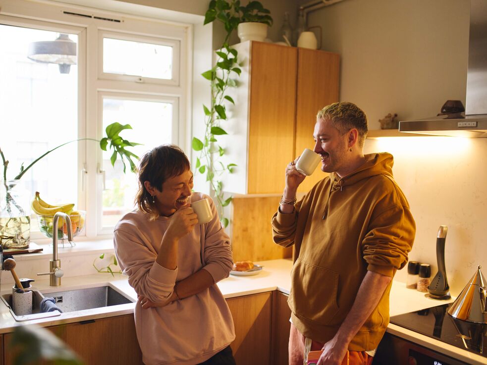 Zwei Personen stehen in einer Küche, trinken aus Tassen und unterhalten sich am Spülbecken.