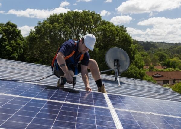 Ein Arbeiter in Schutzkleidung installiert bei teilweise bewölktem Himmel Solarzellen auf einem Dach.