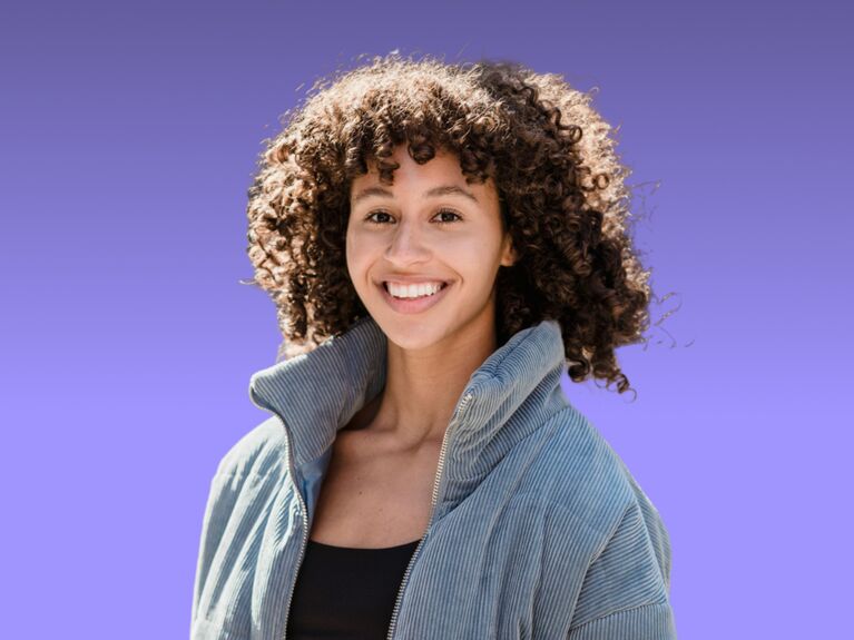 Eine Person mit lockigem Haar lächelt, während sie eine Jacke trägt, vor einem lila Hintergrund.