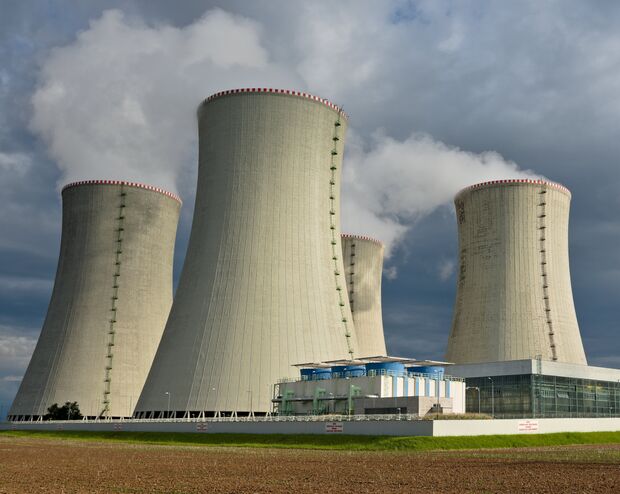Vier große Kühltürme stoßen in der Nähe eines Industriegebäudes bei bewölktem Himmel Dampf aus.