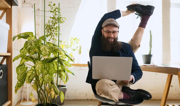 Mann mit Laptop auf einem Stuhl, ein Fuß hinter seinem Kopf