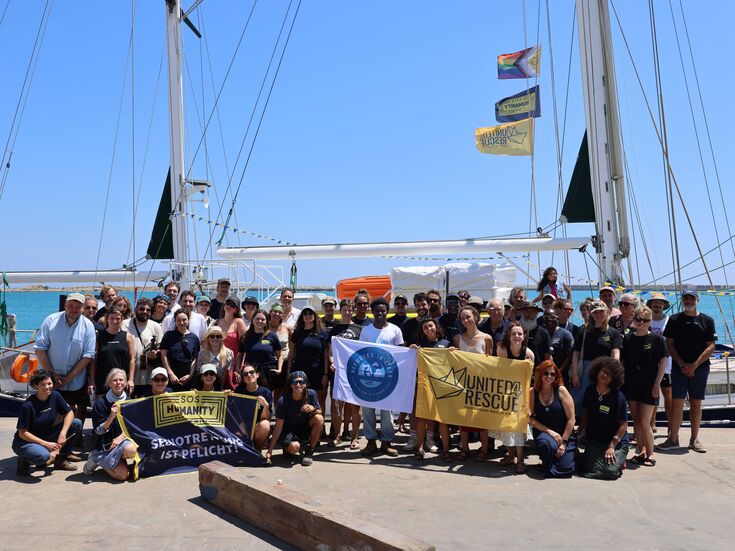 Eine große Gruppe posiert vor einem am Kai liegenden Segelboot und hält dabei Transparente und Fahnen in den Händen.