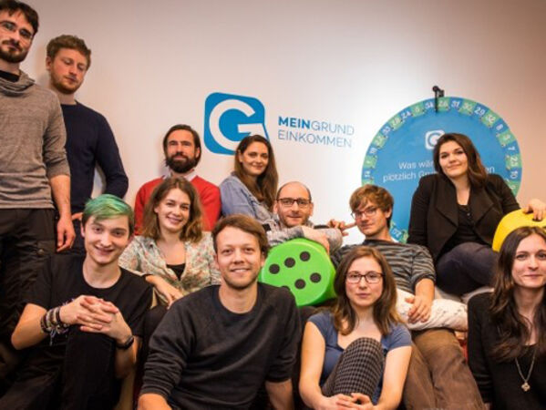 Mitarbeiter*innen der Organisation Mein Grundeinkommen, im Hintergrund ist das Logo des Vereins