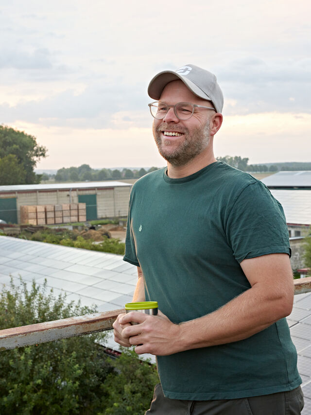 Ein lächelnder Mann mit einer Mütze steht bei Sonnenkollektoren und hält eine grüne Tasse im Freien.