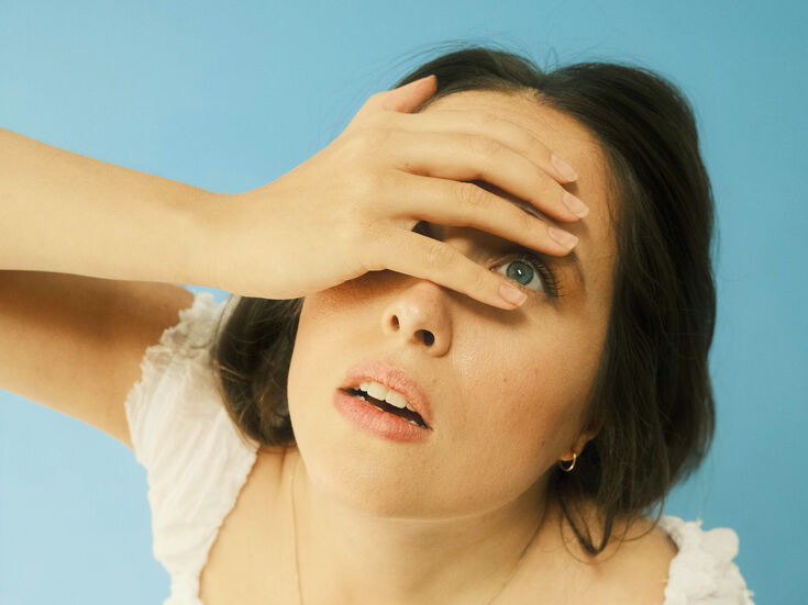 Frau in weißem Oberteil blickt mit einer Hand auf ihre Stirn vor blauem Hintergrund.