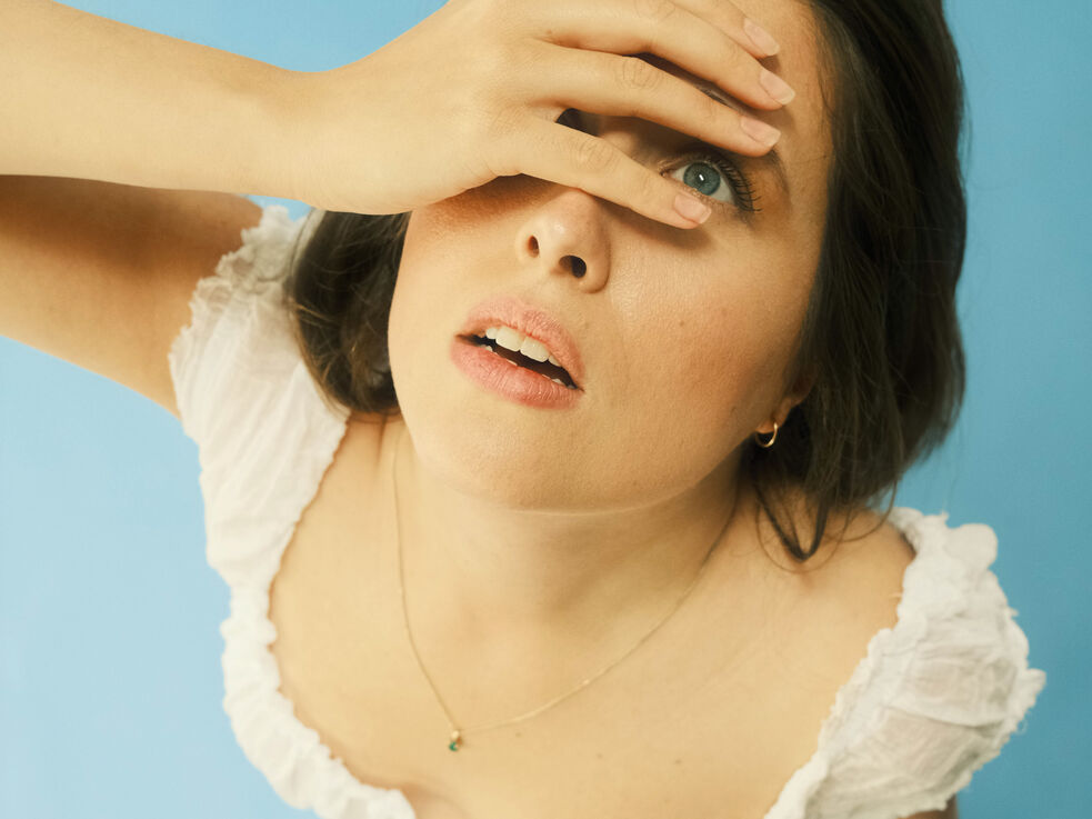 Frau in weißem Oberteil blickt mit einer Hand auf ihre Stirn vor blauem Hintergrund.