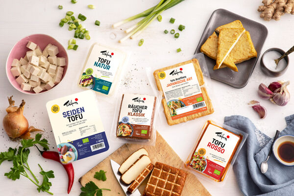 Verschiedene Tofuprodukte mit Gemüse und Kräutern auf einer weißen Tischplatte.