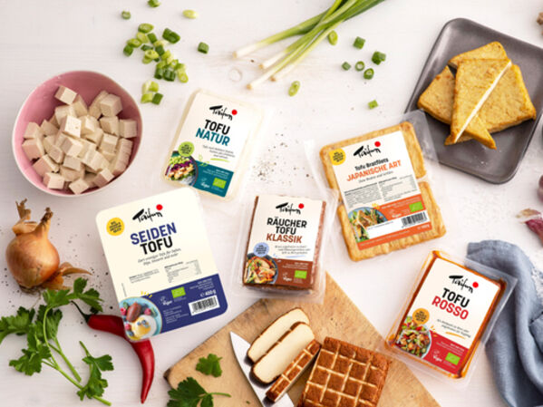 Verschiedene Tofuprodukte mit Gemüse und Kräutern auf einer weißen Tischplatte.
