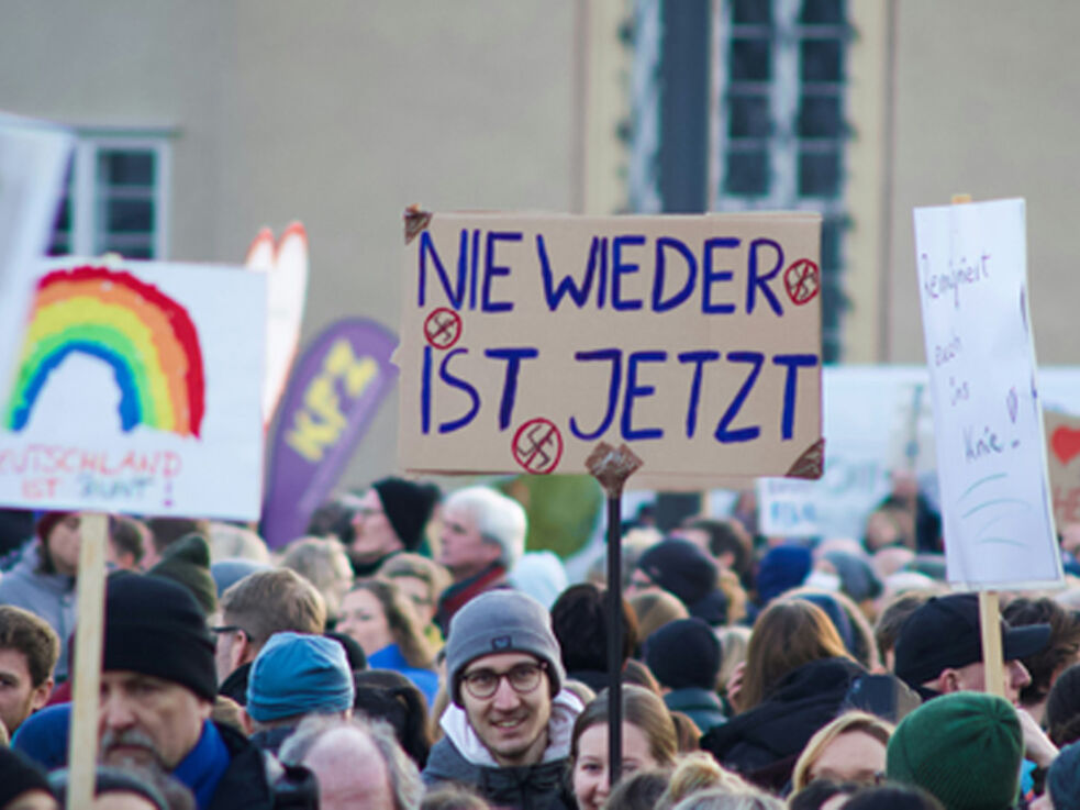 Demonstration: Ein*e Teilnehmer*in hält ein Plakat mit der Aufschrift "Nie wieder ist jetzt" und durchgestrichenen Hakenkreuzen